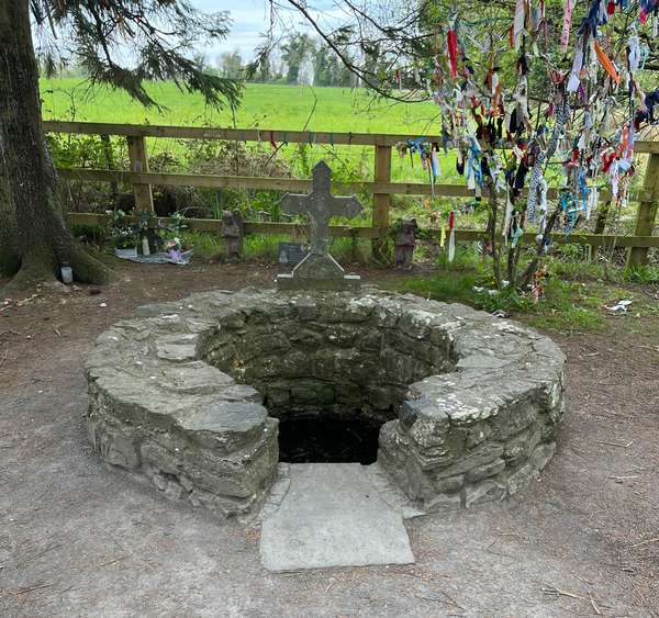 Brigid's well is made out of stacked stones with a cross made of stone. Behind it sits a tree with clooties hanging from it.